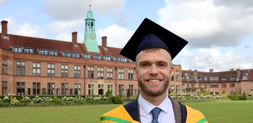 Colin Hartley wearing his graduation cap and gown over a shirt and tie stands on the lawn in front of the HCA Building.